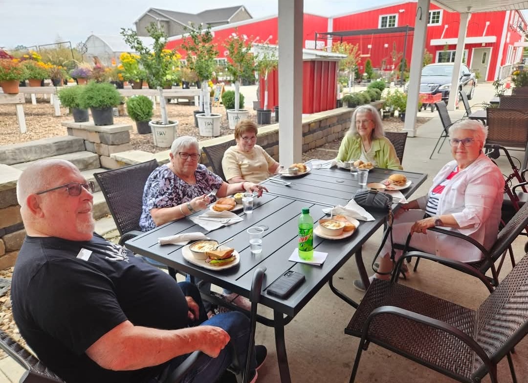 Residents dine outdoors at Blooms Garden Center, surrounded by colorful flowers and a red barn.