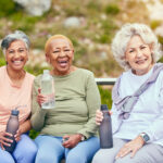 Three older women sitting outdoors in warm weather, smiling and holding water bottles, representing the wellness, connection and comfort of a summer move to senior living in Cedar Rapids, Iowa.