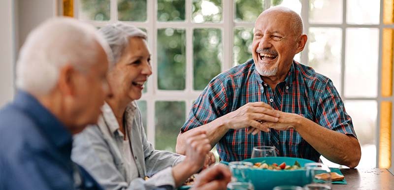 Group of older adults seated together at a table enjoying a meal and talking in a bright dining setting, representing social dining and connection like at Stoney Point Meadows