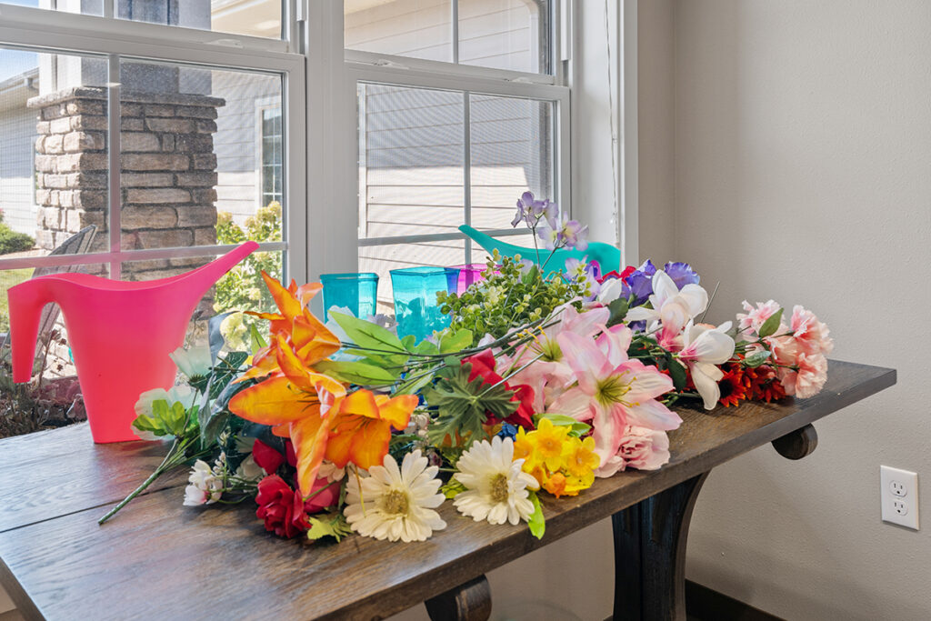 Table with colorful artificial flowers, watering can, and containers near a window, representing craft and activity projects at Stoney Point Meadows