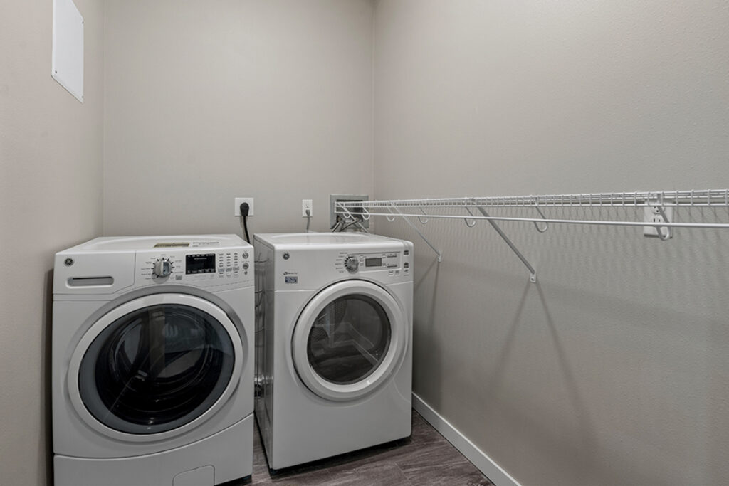Apartment laundry area with a front-loading washer and dryer, wall-mounted shelf, and hanging rod for clothes at Stoney Point Meadows