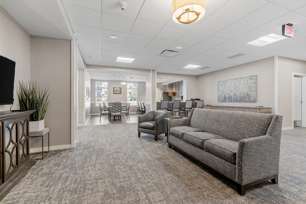 Open lounge area with sofa seating, armchairs, television, and connected dining space in a bright, multi-use common area at Stoney Point Meadows