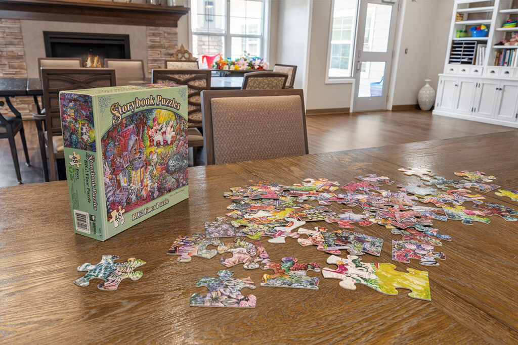 Puzzle pieces spread across a table with a puzzle box nearby in a community activity space with chairs and fireplace at Stoney Point Meadows