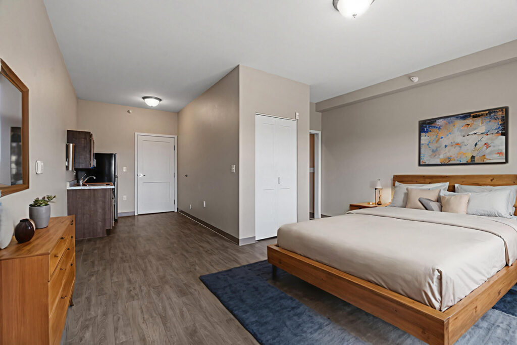 Open studio-style apartment with bed, dresser, kitchenette, closet, and wood flooring in a spacious layout at Stoney Point Meadows