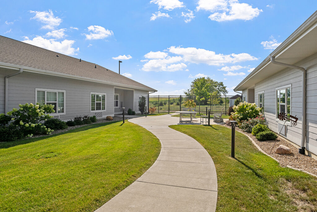 Curved walking path through a landscaped courtyard with lawn, flowers, seating area, and surrounding residential buildings at Stoney Point Meadows