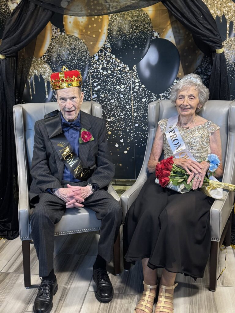 Prom king and prom queen seated in chairs, wearing crowns and sashes, in front of a black-and-gold balloon backdrop. The prom queen holds a bouquet of red roses.