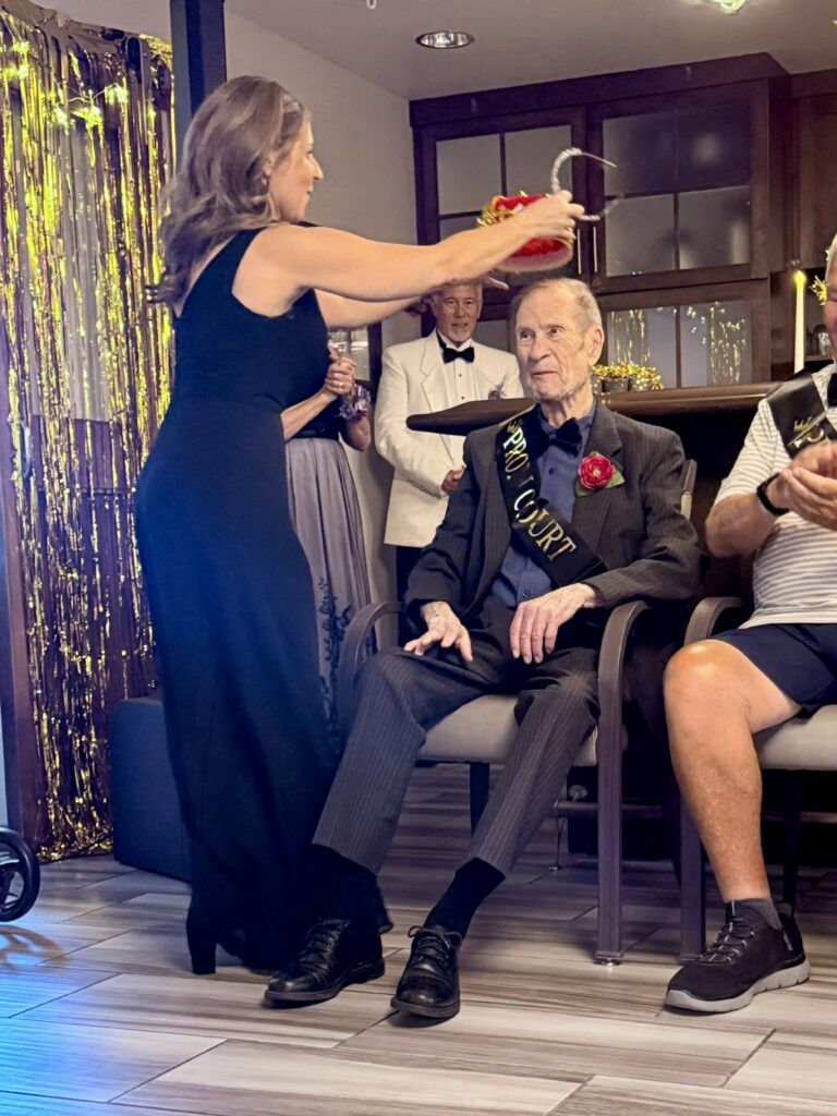 Person places a crown on a seated prom court member wearing a suit, sash, and boutonniere during a prom-themed celebration.