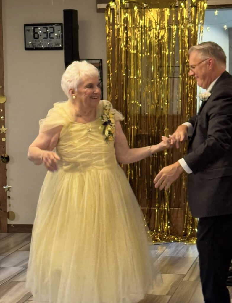 Person in a formal gown dances with a partner in a suit in front of a gold prom-themed backdrop.