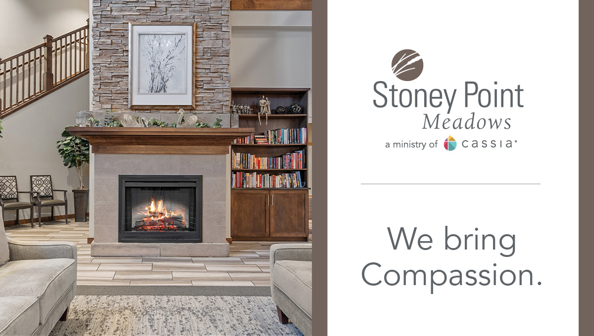 Cozy community fireplace lounge with seating, bookshelves, and a sign that reads “Stoney Point Meadows. We bring Compassion.”